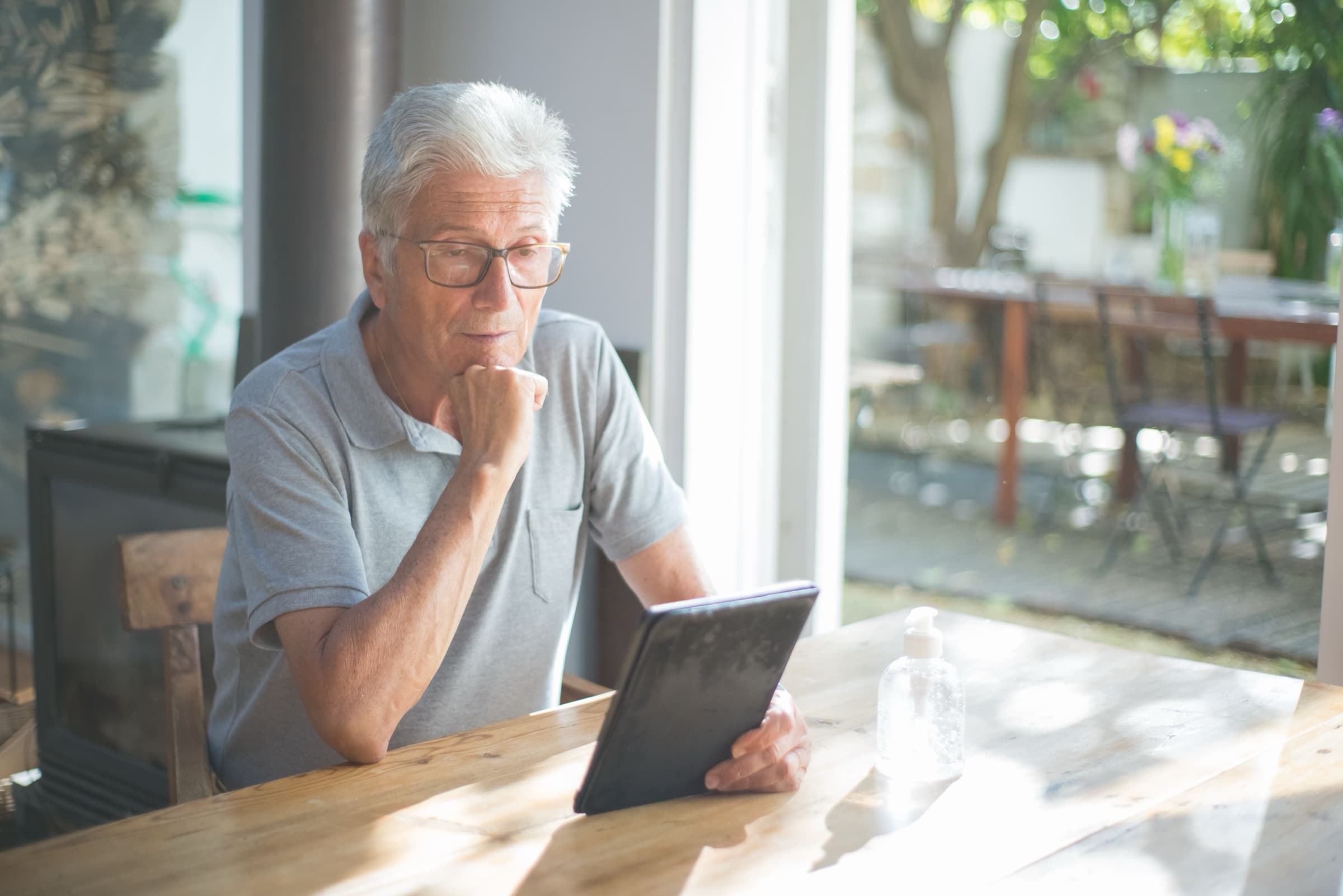 An older man in glasses reads a tablet at a sunlit wooden table at home.